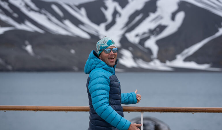 Chris Davenport pictured smiling with a double thumbs up, color blocked jacket, beanie and sunglasses, overlooking the water and a snow capped peak.