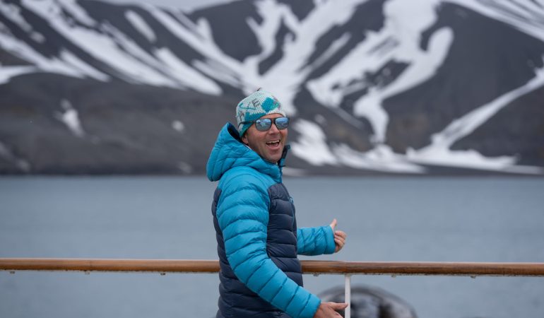 Chris Davenport pictured smiling with a double thumbs up, color blocked jacket, beanie and sunglasses, overlooking the water and a snow capped peak.