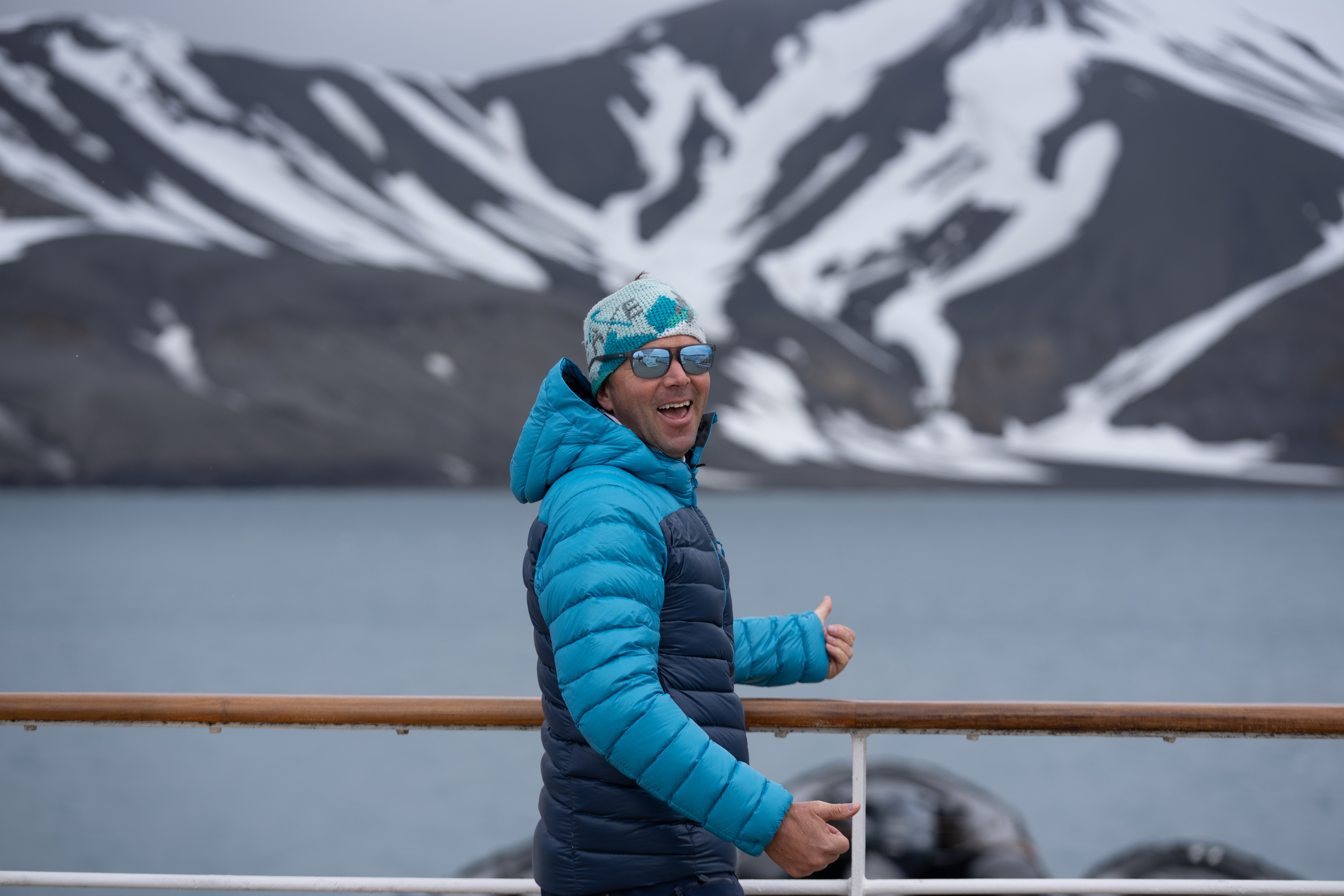 Chris Davenport pictured smiling with a double thumbs up, color blocked jacket, beanie and sunglasses, overlooking the water and a snow capped peak.