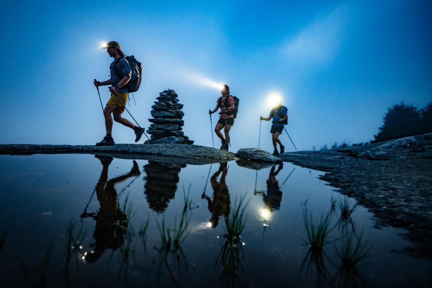 Three hikers at dusk with headlights. Their reflections show in the water and they are silhouettes against the sky.