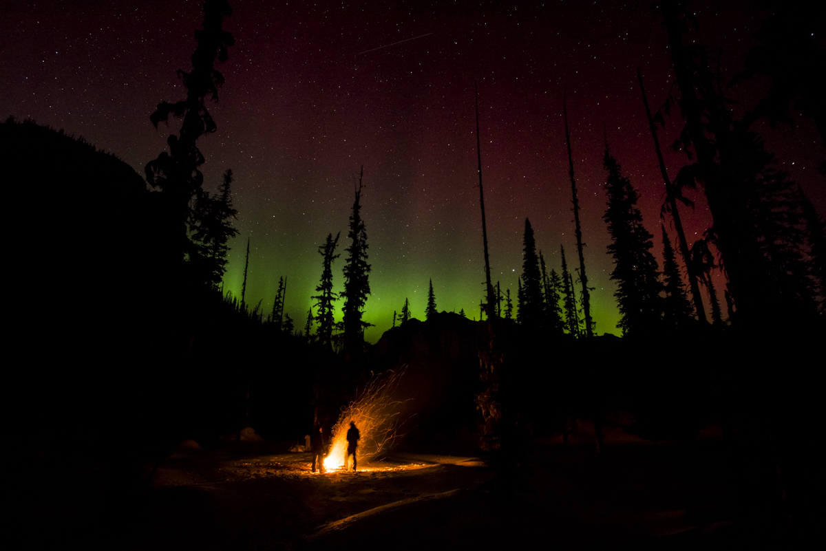 Campfire at night with the black outline of trees against a stunning green and red night sky