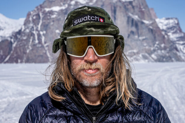 Photo of Cody Townsend in a beanie and goggles with his hair down in front of a snowy peak.