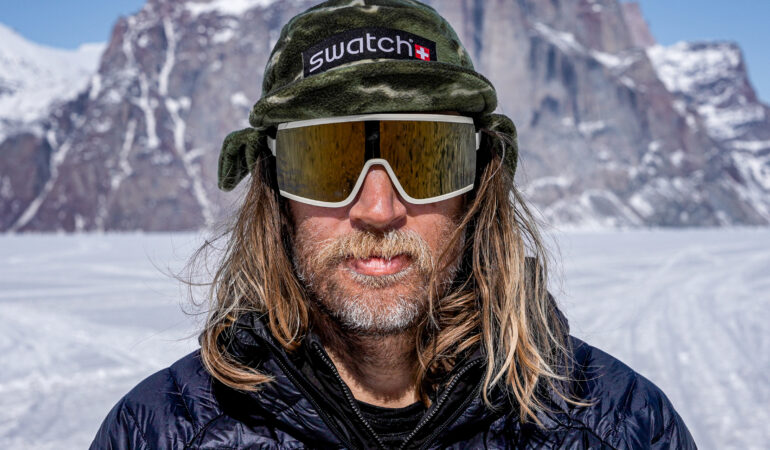 Photo of Cody Townsend in a beanie and goggles with his hair down in front of a snowy peak.