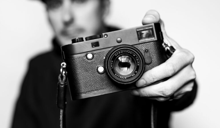 Blurred black and white background of man holding a camera, all focus centering on the camera in his hands