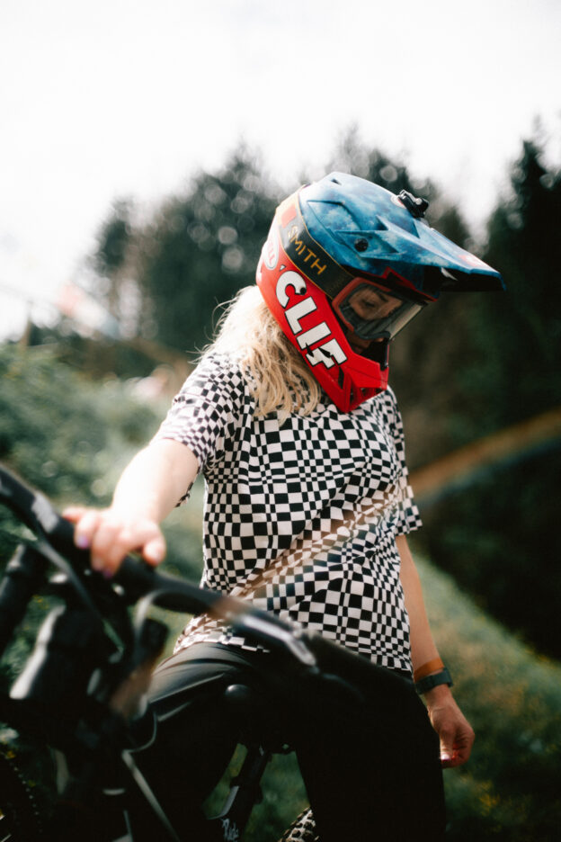 Photo of Casey Brown posing with her mountain bike. Light leaks overlay a black and white checkered shirt, and her face is covered with a heavy CLIF branded helmet and goggles.