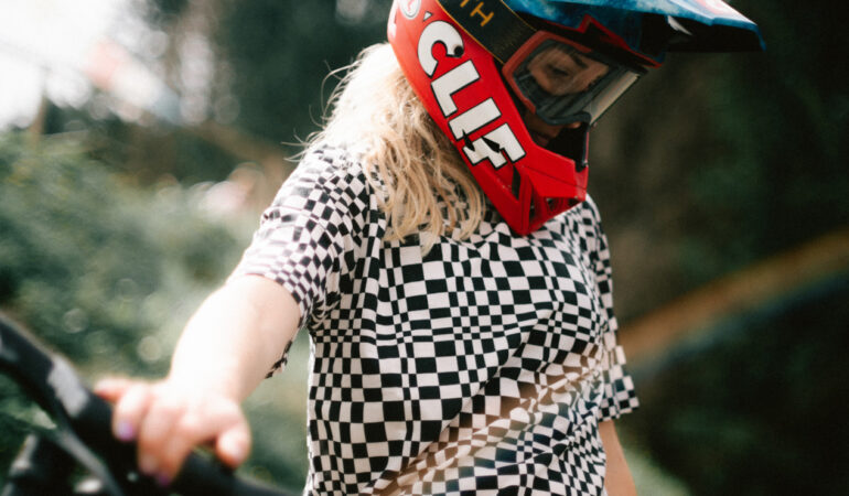 Photo of Casey Brown posing with her mountain bike. Light leaks overlay a black and white checkered shirt, and her face is covered with a heavy CLIF branded helmet and goggles.