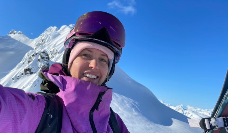 Selfie of Ingrid in purple gear, goggles and a snowy mountain backdrop, smiling at the camera.