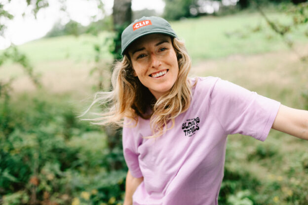 Casey Brown in a lavender shirt and green CLIF baseball cap. She is smiling at the camera in front of a lush green outdoor backdrop.