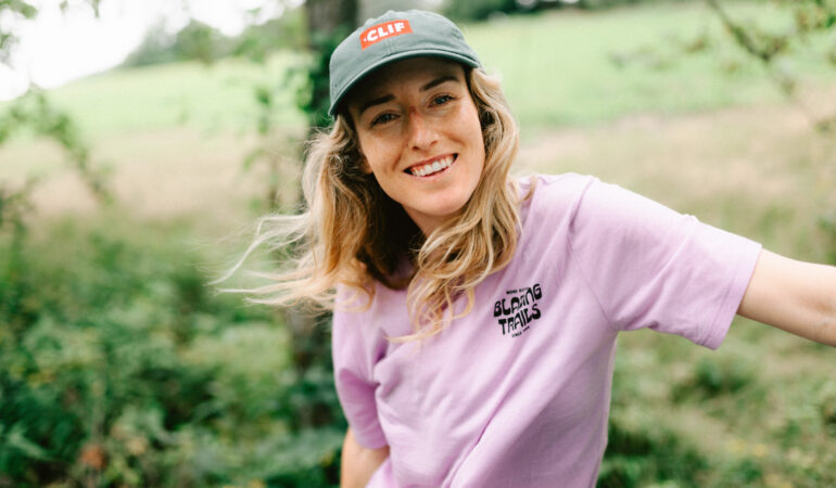 Casey Brown in a lavender shirt and green CLIF baseball cap. She is smiling at the camera in front of a lush green outdoor backdrop.