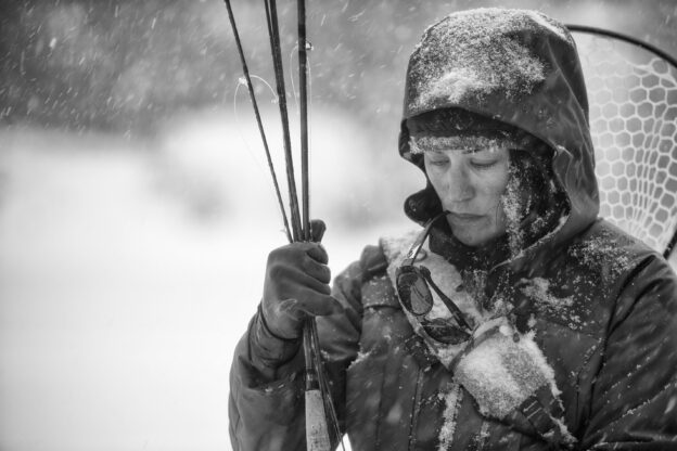 Black and white image of Hilary in the snow, holding her fly fishing gear and sunglasses dangling from her mouth.