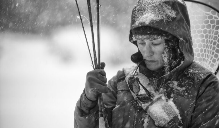 Black and white image of Hilary in the snow, holding her fly fishing gear and sunglasses dangling from her mouth.